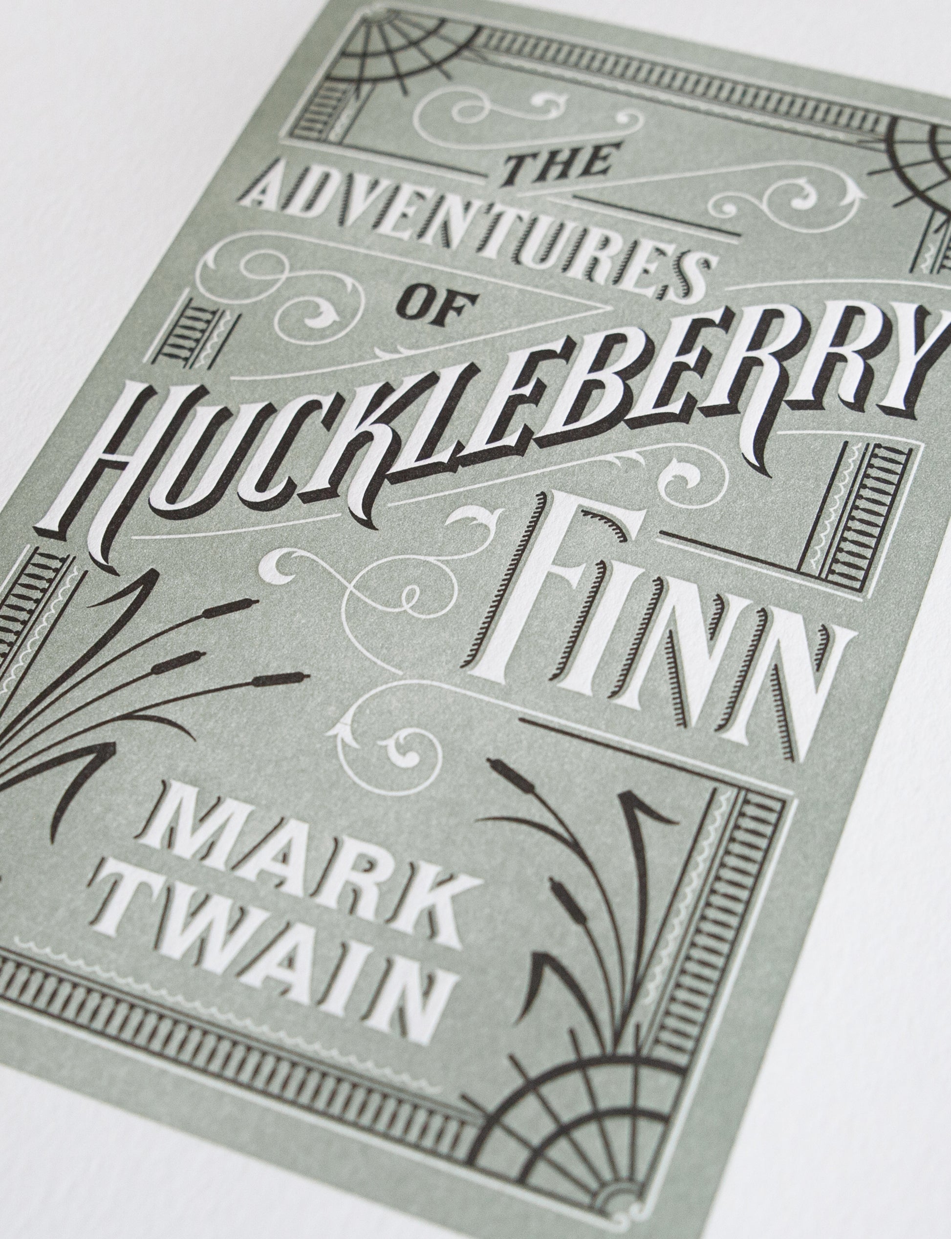 Close-up of a 2-color letterpress print in green and black. Printed artwork is an illustrated book cover of The Adventures of Huckleberry Finn including custom hand lettering.